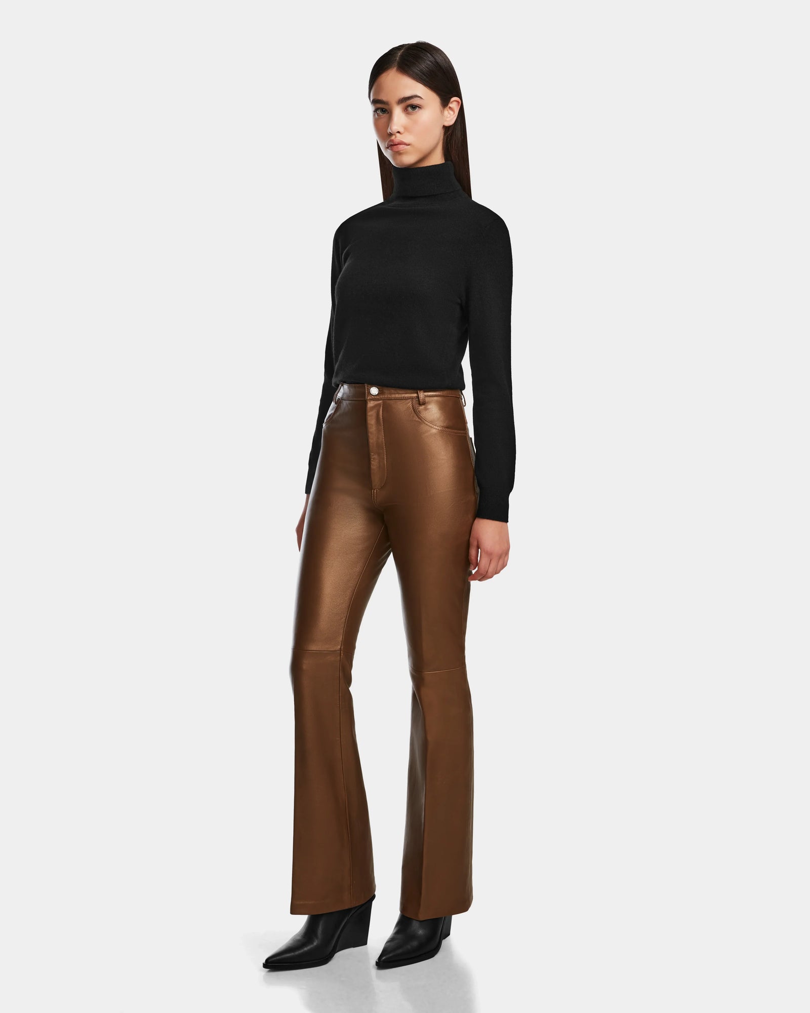 A woman with straight brown hair and a neutral expression stands against a plain background, wearing the Rudsak CARISSA - 8524545 Antique Bronze high-waisted women’s leather pants, a black turtleneck sweater, and black boots.