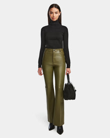 A woman in a black turtleneck and Rudsaks CARISSA - 8524545 Loden Green flare leather pants stands facing forward, holding a black handbag in her left hand with black pointed-toe shoes against a plain white background.