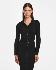 A woman with straight brown hair models the Rudsak CARLI - 8524573 Black, a fitted long-sleeve knit dress with front buttons and a narrow vegan leather belt, standing against a plain white background.