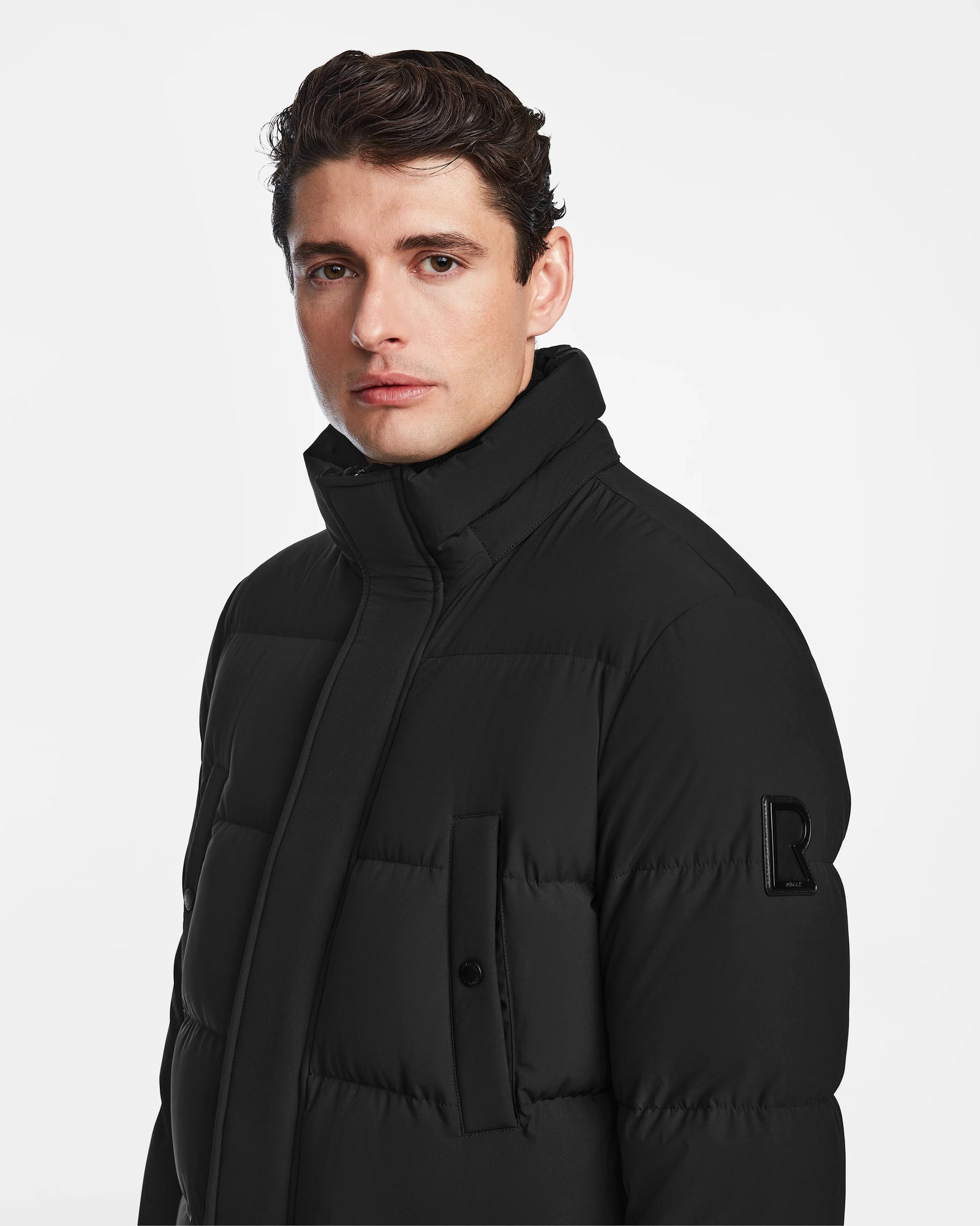 A man with short dark hair models the Rudsak CARLOS - M124509 Black men’s down parka with a high collar and “B” patch on the upper sleeve, standing against a plain white background and glancing to the side.