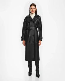 A woman stands facing forward in the Rudsak CASSIE L - 8124531 Black leather trench coat, paired with black knee-high boots and a white turtleneck sweater against a plain white background.