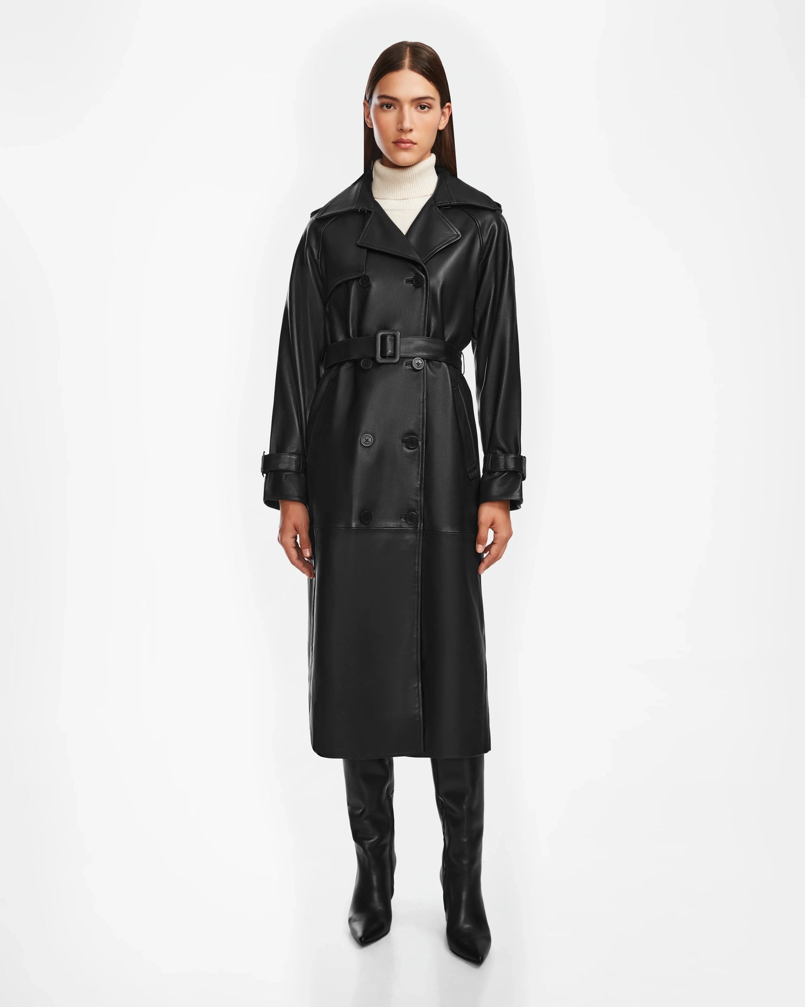 A woman stands facing forward in the Rudsak CASSIE L - 8124531 Black leather trench coat, paired with black knee-high boots and a white turtleneck sweater against a plain white background.