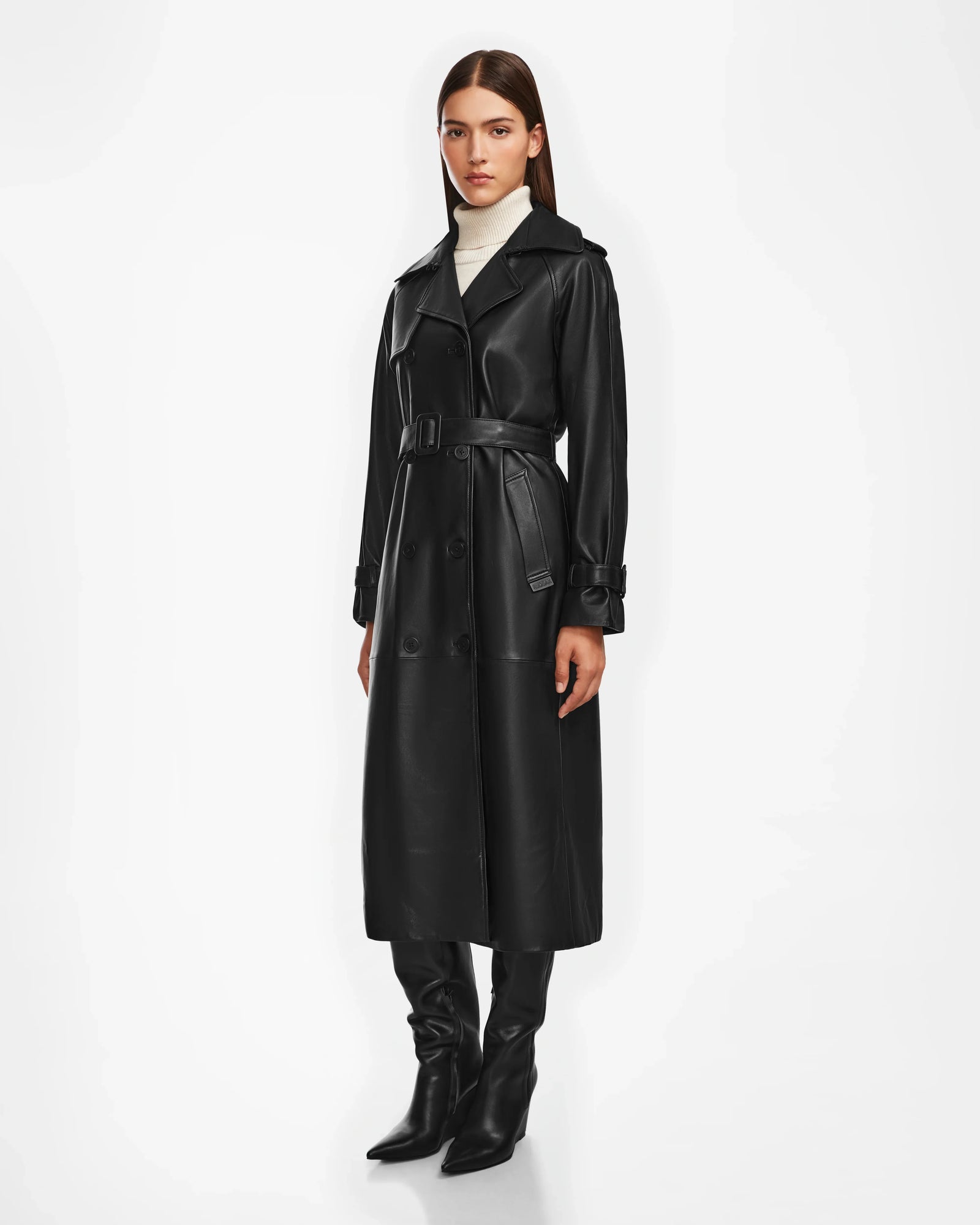 A woman faces forward against a white background, wearing the Rudsak CASSIE L - 8124531 Black trench coat, black knee-high boots, and a white turtleneck.