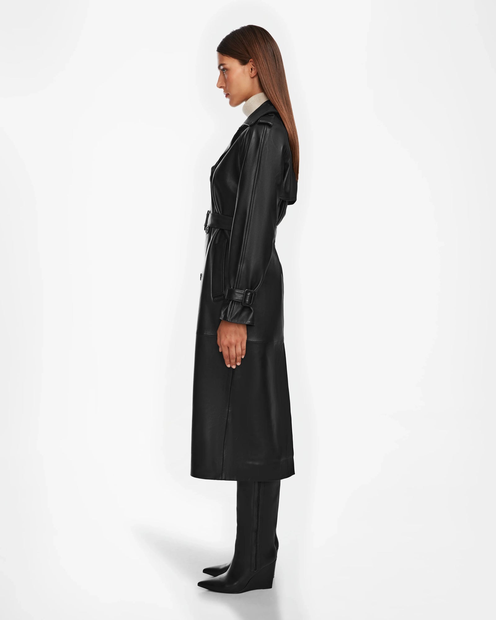 A woman stands in profile wearing the Rudsak CASSIE L - 8124531 Black leather trench coat with high-heeled boots and a light-colored turtleneck, set against a plain white background.