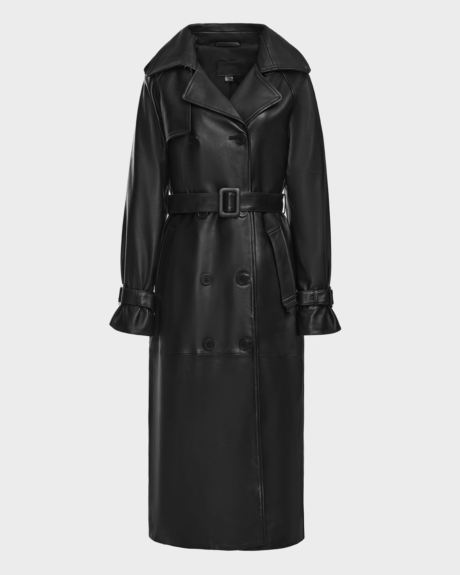 The Rudsak CASSIE L - 8124531 is a women’s long black faux leather trench coat with a wide collar, belted waist, double-breasted buttons, and adjustable cuff straps, shown against a plain white background.