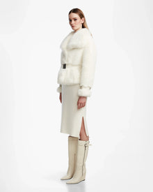 A woman stands sideways wearing the Rudsak CHLOE - L124564 Iceland womens faux fur jacket with belt, a cream midi skirt with side slit, and knee-high vegan leather boots against a plain white background—effortless winter elegance.