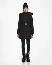 A person stands facing forward wearing the Rudsak CHRISTEL M-M - L124505 Black quilted winter coat with a belt, paired with black leggings, boots, and a pom-pom knit hat, against a plain white background.