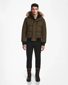 A man stands facing forward, wearing the Rudsak DALLAS - M122501 Army weather-resistant puffer jacket with a fur-trimmed hood, black pants, and brown lace-up boots against a plain white background.