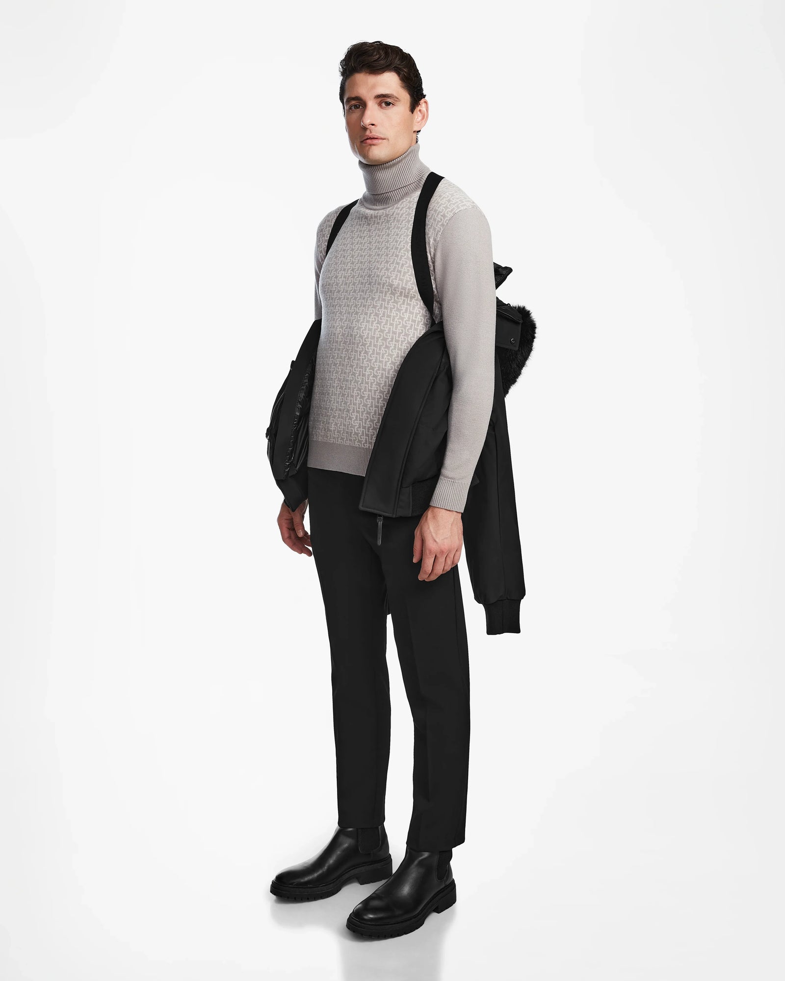 A man poses against a white background in black pants, black boots, and a gray turtleneck, carrying the Rudsak DALLAS - M122501 Black weather-resistant jacket draped over his shoulder as he gazes slightly to the side.