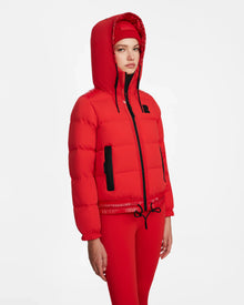 A woman wears the Rudsak DANI M - 8124539 bomber-style ski jacket in Flame Red with a hood, red leggings, and a textured red beanie, standing against a plain white background.
