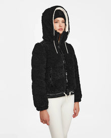 A person wears the Rudsak DANI S - 8124537 Black bomber-style ski jacket with cream drawstrings, white leggings, and a black beanie, standing against a plain white background.