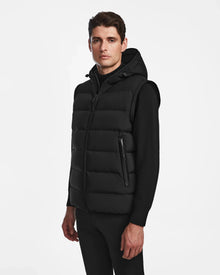 A man wears the Rudsak DEMIAN M-R - M123504 Black down puffer hooded vest over a black long-sleeve shirt and pants, standing against a white background with a neutral expression.