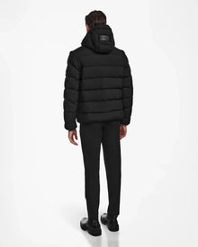 A man faces away, wearing the Rudsak DEMIAN M-R - M123504 Black mens down puffer made of recycled matte fabric with a hood, paired with black pants and shoes, set against a plain white background.