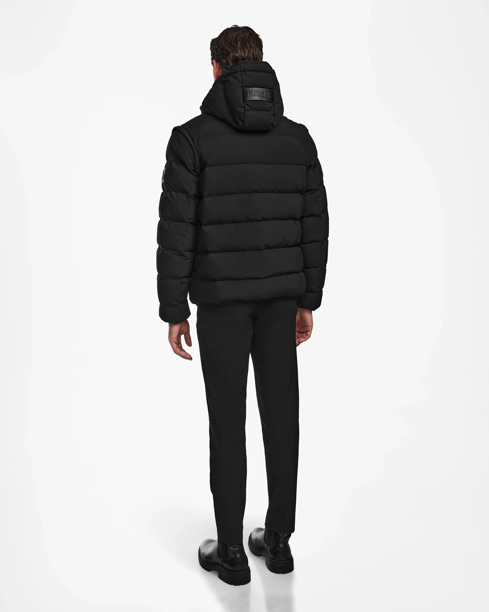 A man faces away, wearing the Rudsak DEMIAN M-R - M123504 Black mens down puffer made of recycled matte fabric with a hood, paired with black pants and shoes, set against a plain white background.