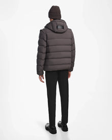 A person stands facing away, wearing the Rudsak DEMIAN M-R - M123504 Grey men’s down puffer jacket with a hood, paired with black pants, black shoes, and a brown knit beanie—all set against a plain white background.