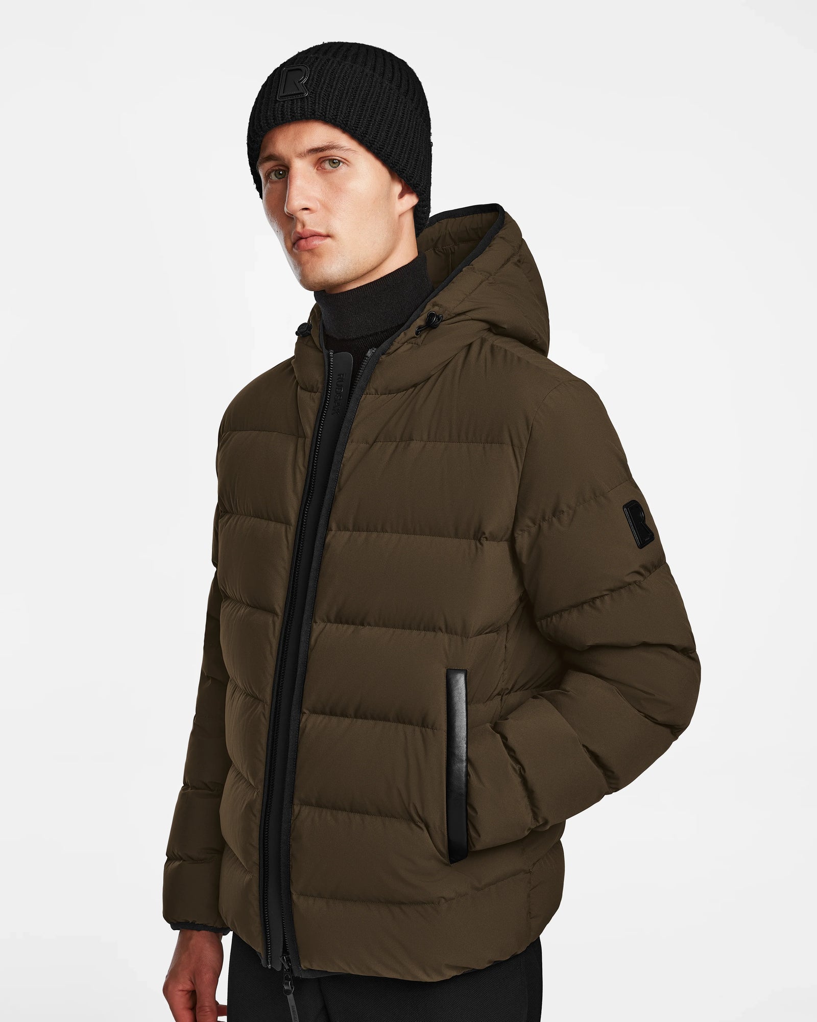 A man wearing a black beanie, black turtleneck, and the Rudsak DEMIAN M - M123507 Army mens puffer jacket stands against a plain white background.