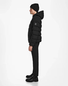 A man stands in profile against a white background, wearing the Rudsak DEMIAN M - M123507 Black hooded down puffer, paired with a black knit beanie, black pants, and black boots.