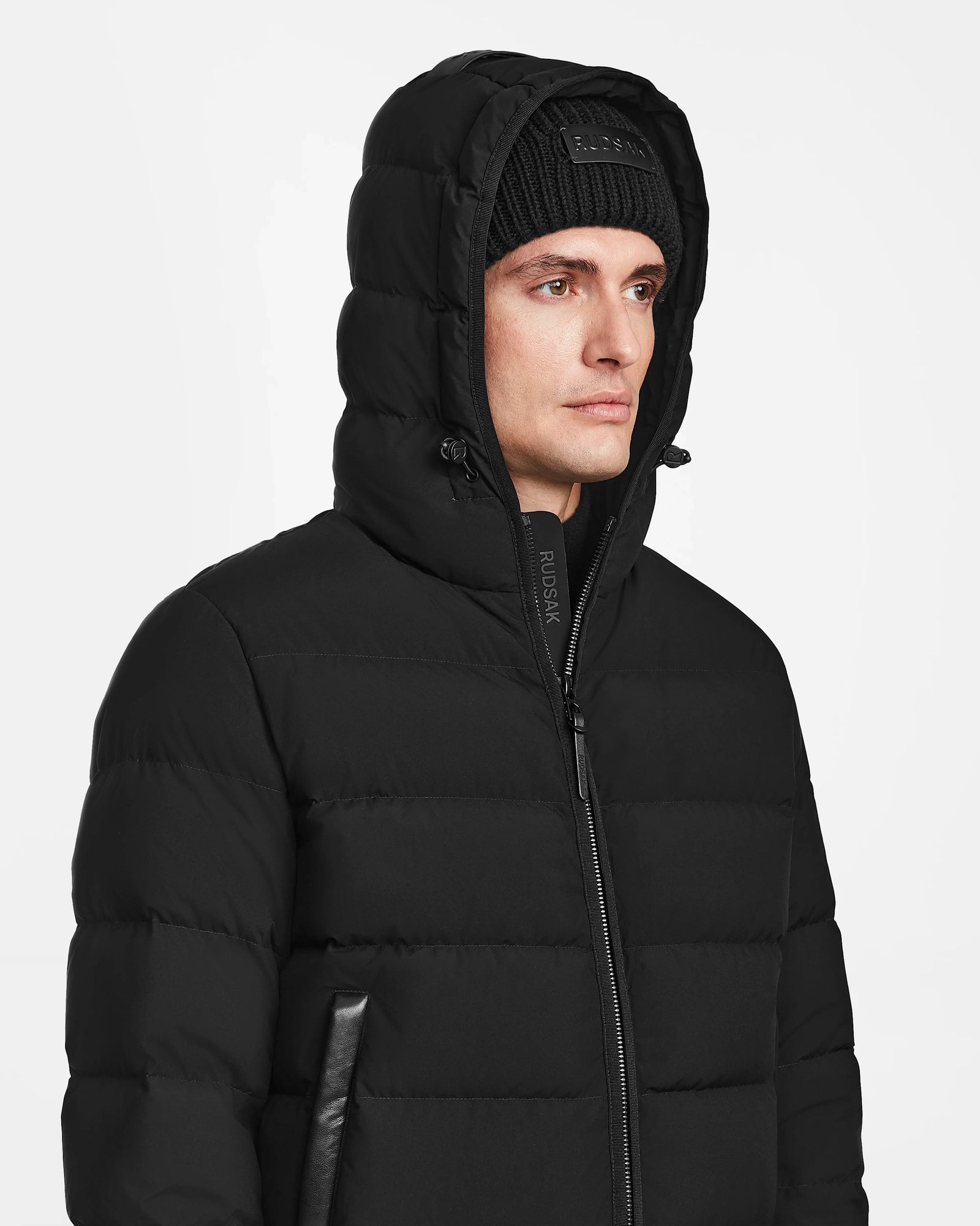 A man wearing the Rudsak DEMIAN M - M123507 Black hooded down puffer, crafted from sustainable recycled fabric, with the hood up and a black knit beanie, stands against a plain white background.