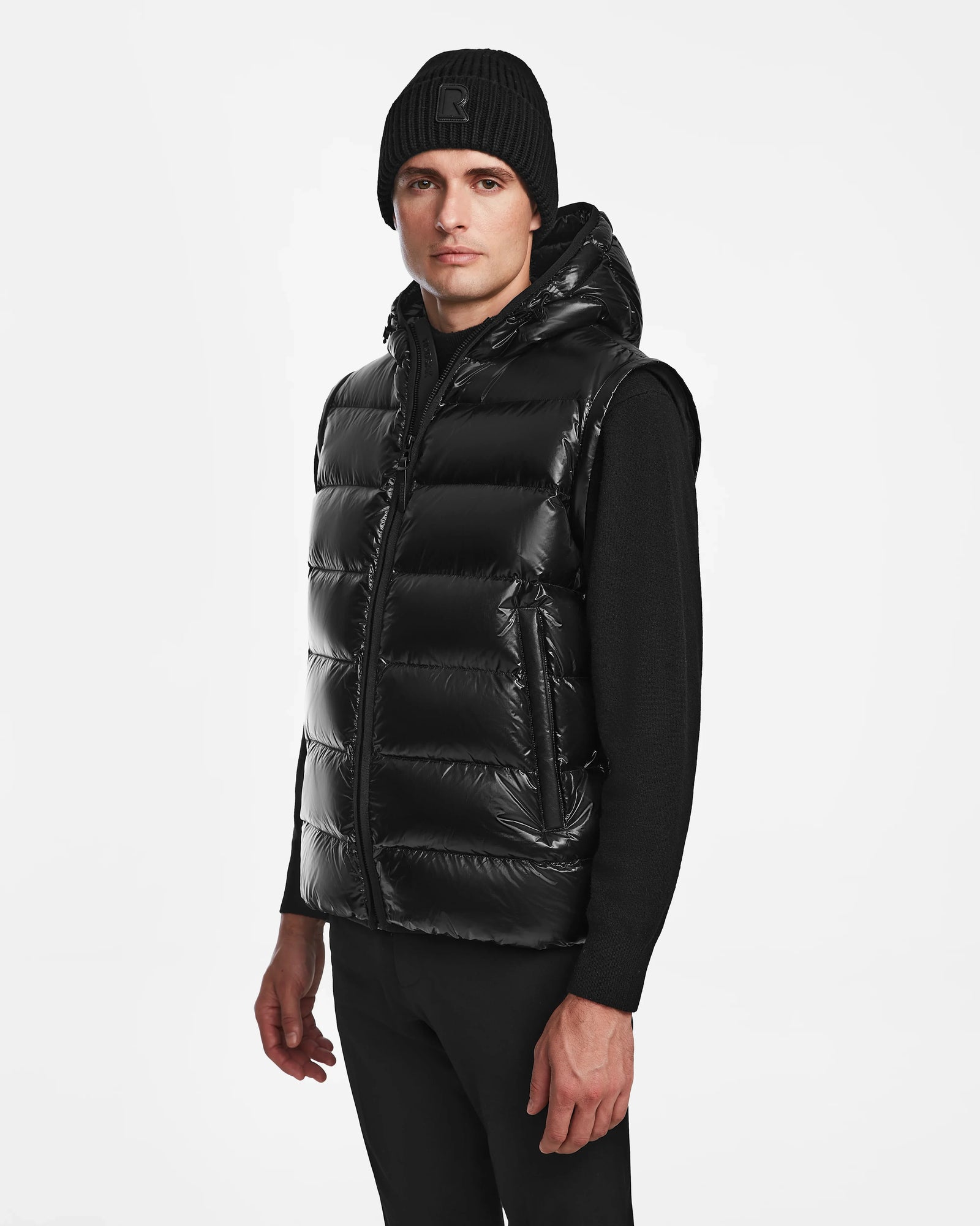 A man wearing Rudsaks DEMIAN S-R - M123505 Black down puffer vest with a hood, styled with a black beanie, long-sleeve shirt, and pants, stands against a plain white background.