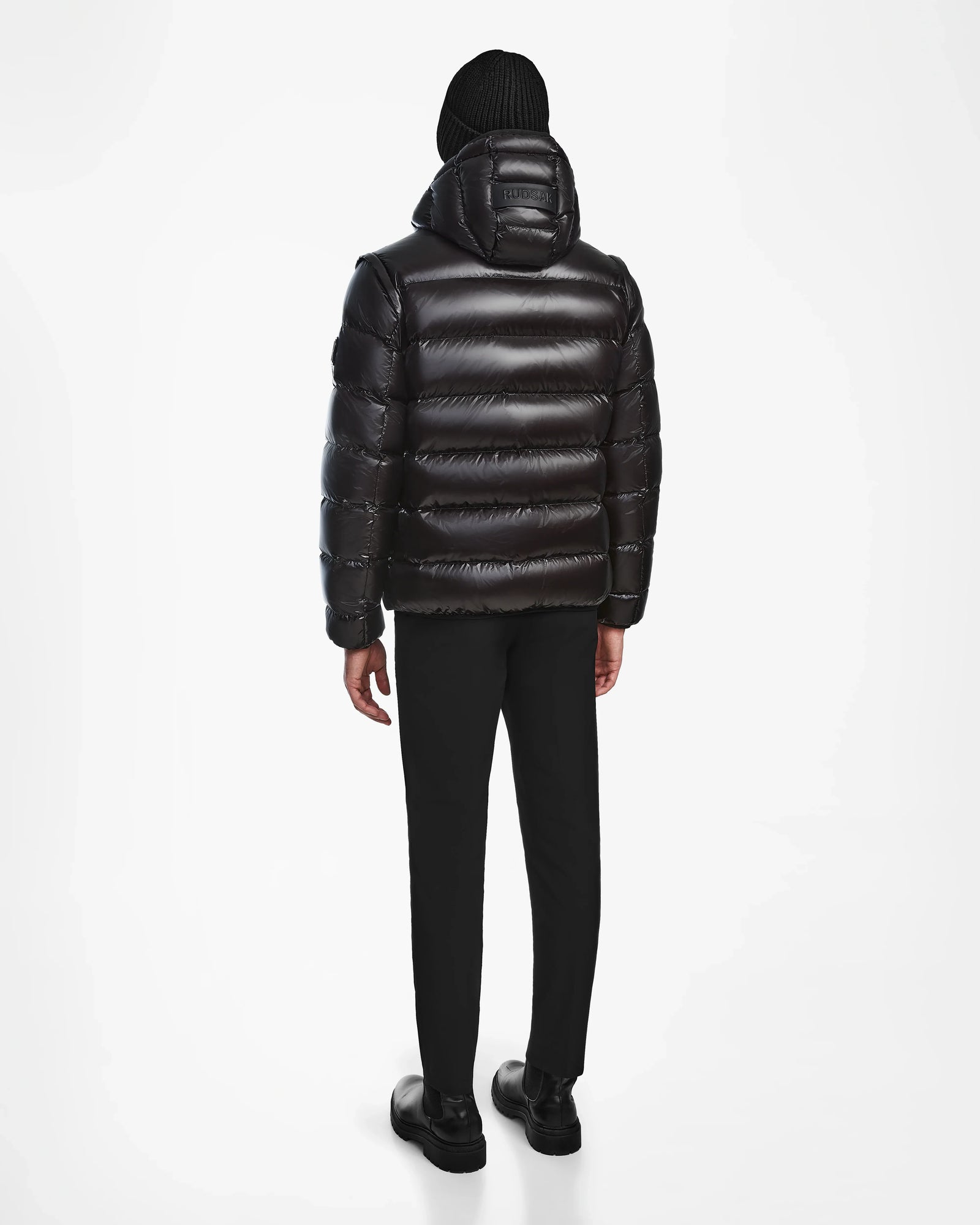 A person faces away against a white background, wearing the Rudsak DEMIAN S-R - M123505 Black mens down puffer jacket with hood, paired with black pants, shoes, and a black knit beanie.