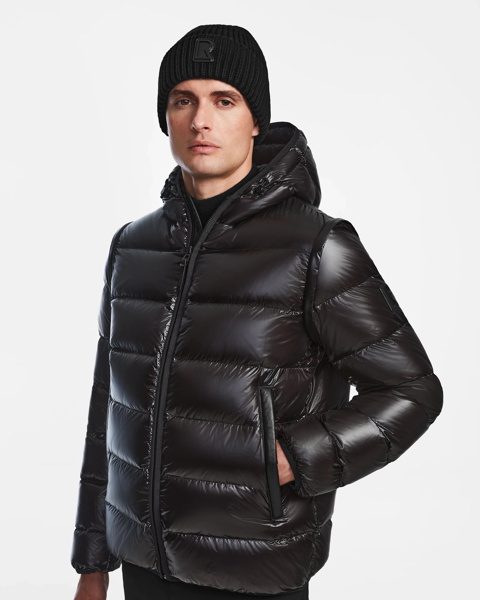 A person wears the Rudsak DEMIAN S-R - M123505 Black mens down puffer jacket with a hood and a black knit beanie, standing against a plain white background. This RDS-certified jacket delivers premium warmth and sleek style.