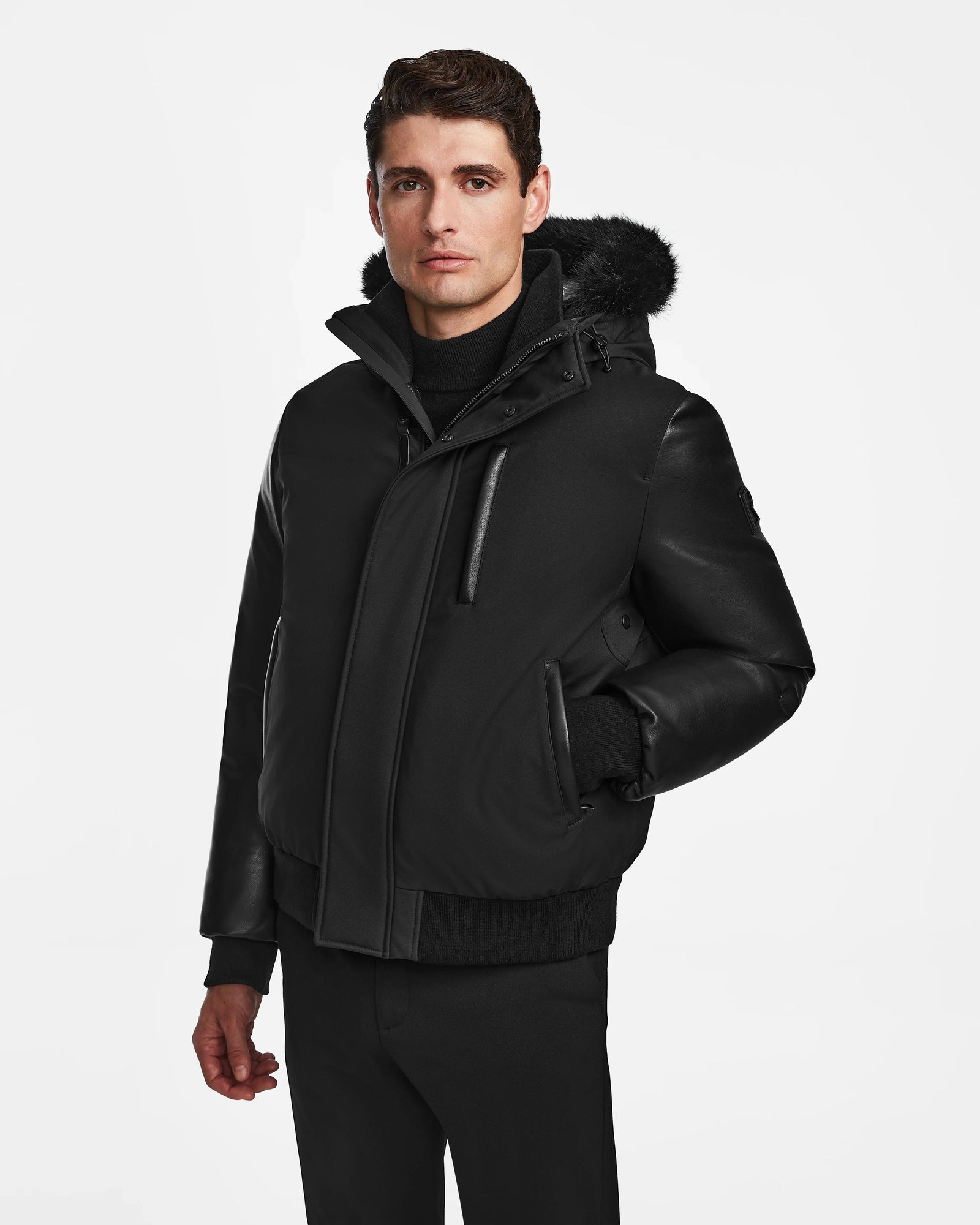 A man in the Rudsak DERON - M123517 BLACK mens bomber jacket, featuring a fur-lined hood, stands against a white background with one hand in his pocket. This stylish weather-resistant piece is perfect for chilly days.