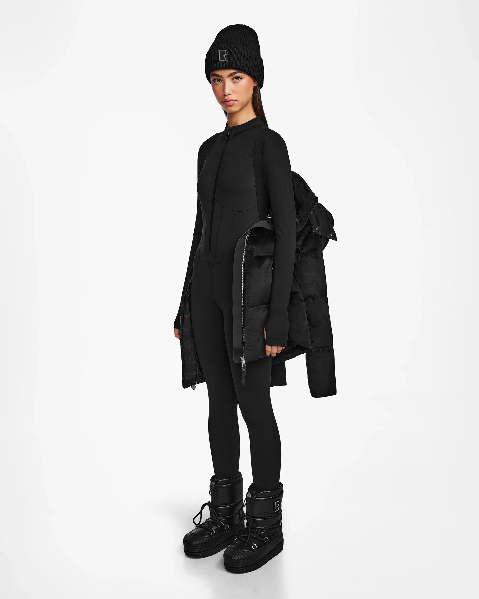 A woman in the Rudsak DONIA - L124563 Black down puffer, black beanie, fitted jumpsuit, and black winter boots stands against a white background.