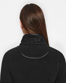 A person with straight brown hair is seen from behind, wearing a black Rudsak EDITH - 8524617 womens zip-up sweater with the brands name embroidered on the collar.