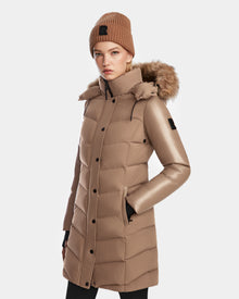 A woman in the Rudsak ELSA - L123535 Sand quilted down puffer with faux fur–trimmed hood and matching knit beanie stands with her hands in her pockets against a plain white background.