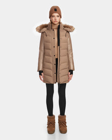 A person stands facing forward, wearing the Rudsak ELSA - L123535 Sand women’s quilted down puffer coat with fur-trimmed hood, a matching knit beanie, tan sweater, black leggings, and tan boots against a plain white background.
