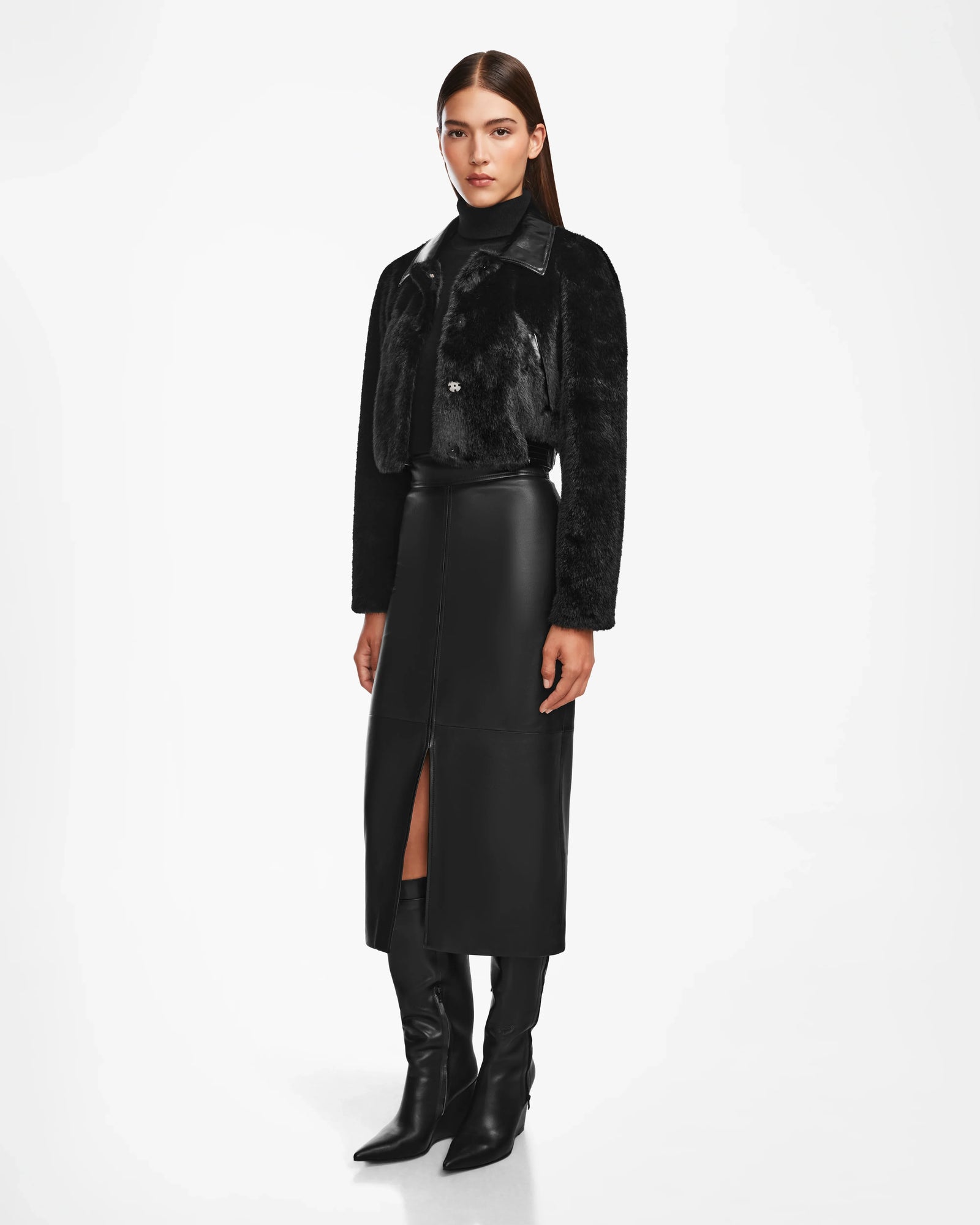 A woman poses against a white background wearing the Rudsak EVELYN - 8124791 Black faux fur jacket, a black leather midi skirt with a front slit, a black top, and pointed black knee-high boots.