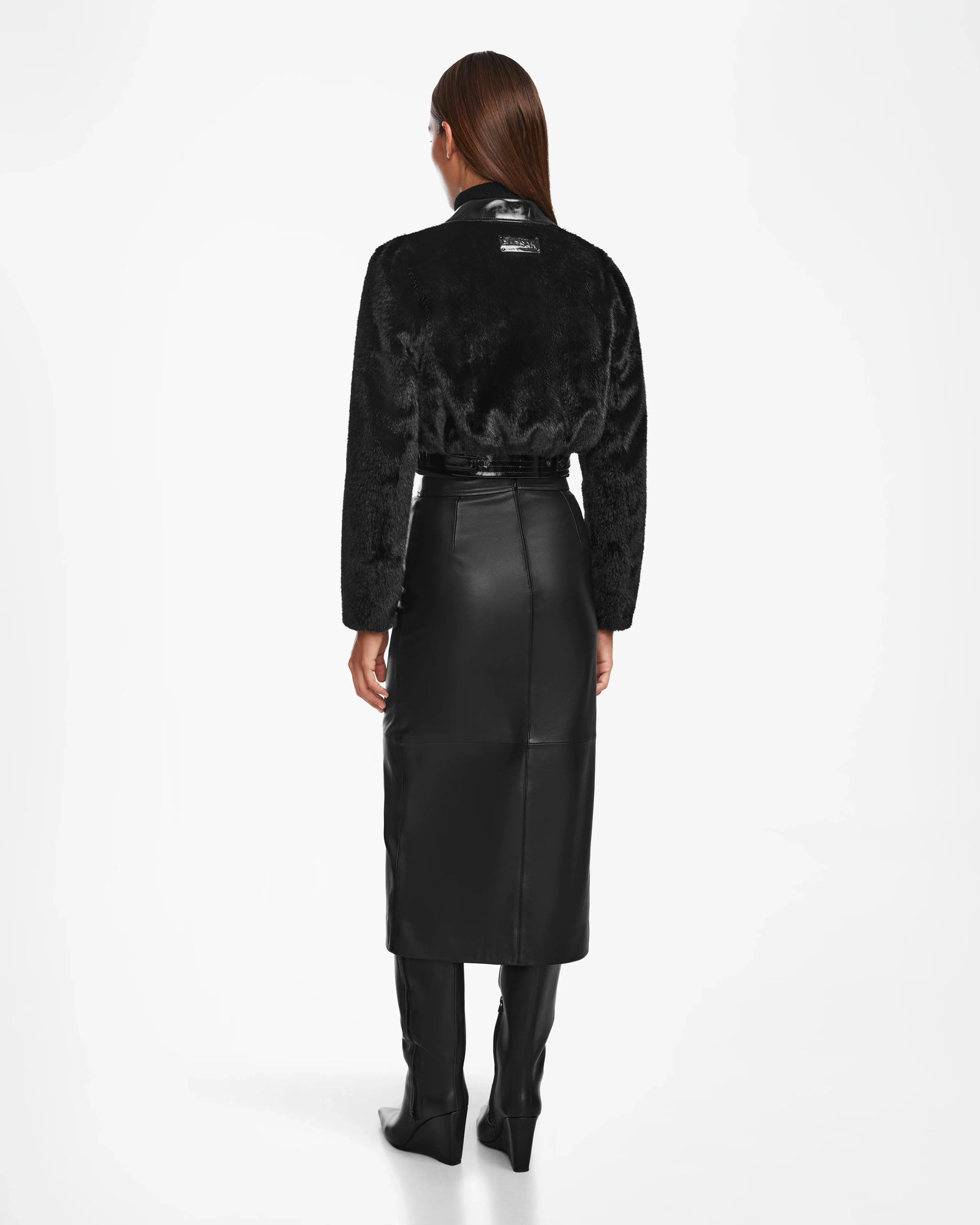 A woman faces away against a white background, wearing the Rudsak EVELYN - 8124791 Black faux fur jacket, paired with a black belted leather skirt below the knees and black knee-high boots.