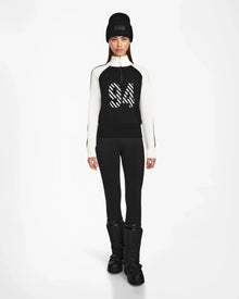 A woman faces forward against a white background, dressed in Rudsak’s FANNY - 8524506 Black après-ski sweater with “84” on the front, black leggings, snow boots, and a knit beanie—ideal for winter layering.