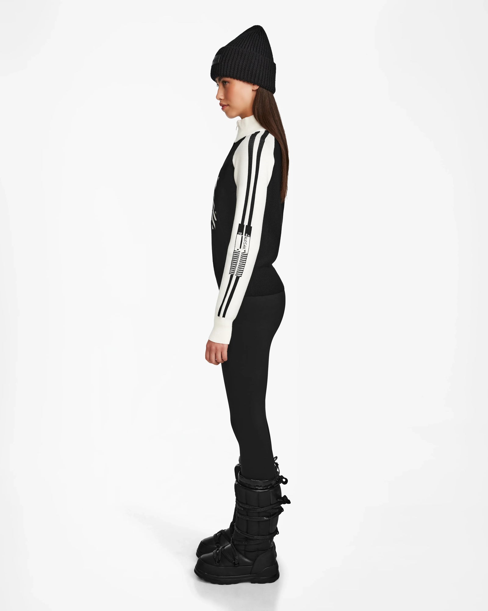 A woman in profile faces left, wearing a black beanie, black leggings, black winter boots, and the Rudsak FANNY - 8524506 Black womens knit pullover, layered perfectly for winter against a plain white background.