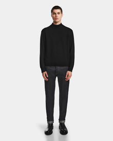 A young man faces forward wearing the Rudsak FERRAN - 6524511 Black mens turtleneck sweater, styled with dark rolled-cuff jeans and black boots against a plain light background.