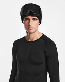 A man wearing the Rudsak FRANC - 6524541 Black stretch base layer top, a black beanie, and black ski goggles on his forehead stands against a plain white background.