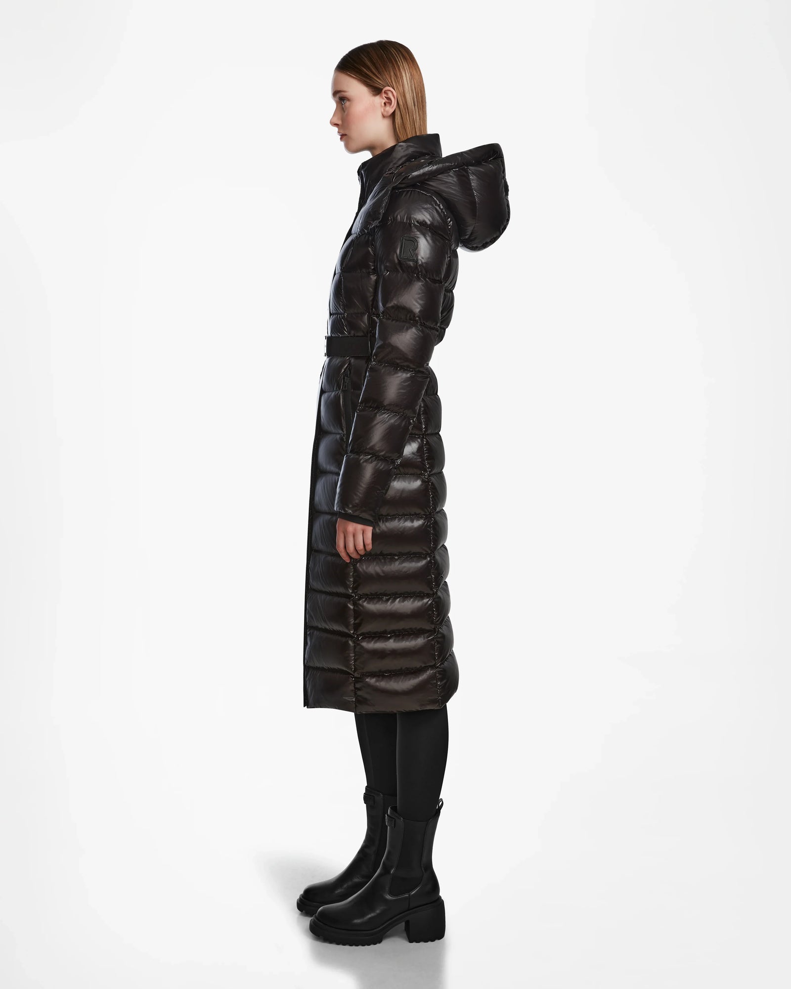 A woman in profile models the Rudsak FRIDA L-S - L124525 Black, a shiny quilted maxi down puffer with hood, paired with black leggings and chunky boots—a chic winter statement—against a plain white background.