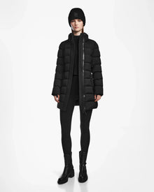 A person stands facing forward against a plain white background wearing the Rudsak FRIDA M - L124524 Black womens mid-length down jacket with a removable hood, paired with a black beanie, black sweater, leggings, and boots.