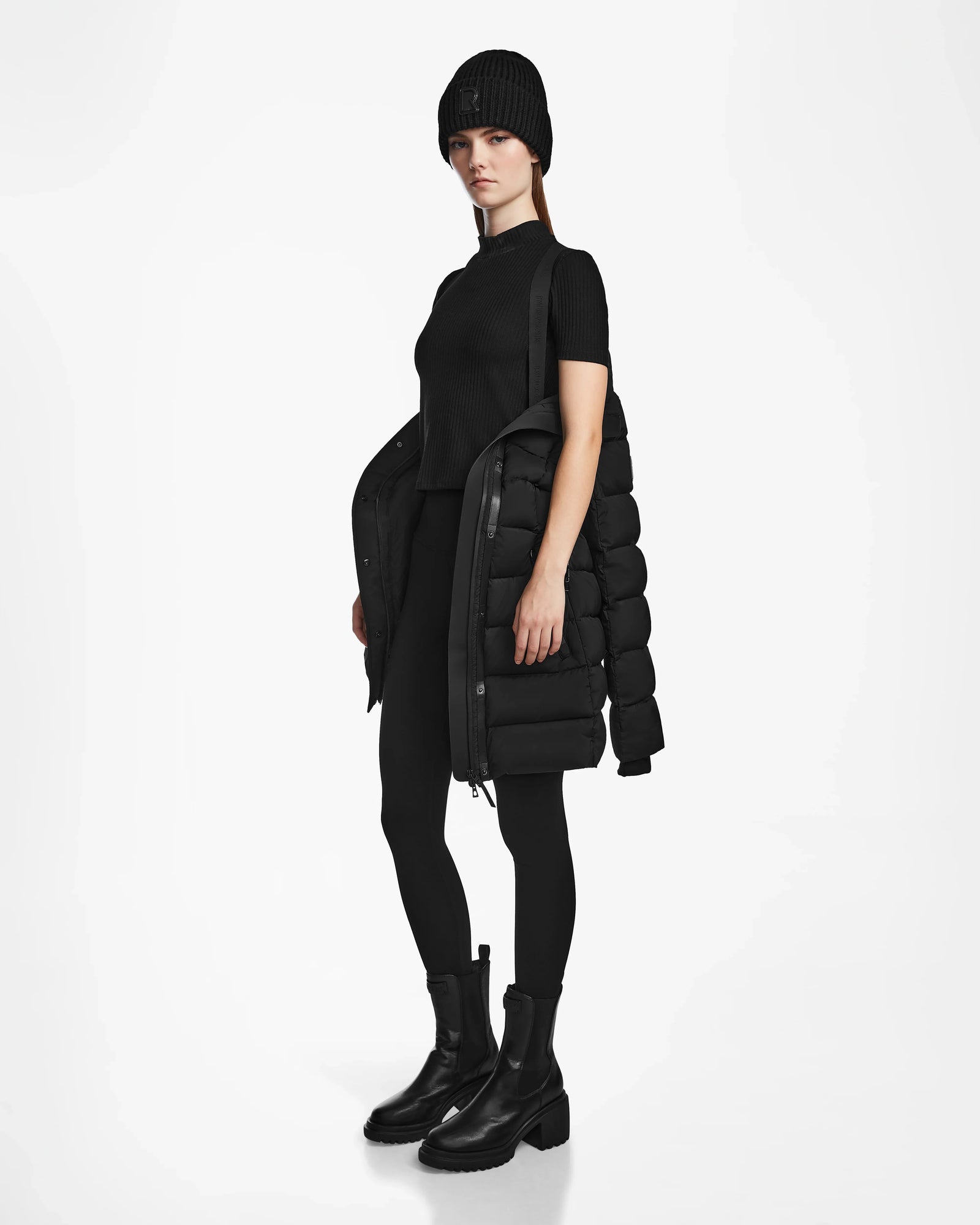 A woman stands against a white background wearing the Rudsak FRIDA M - L124524 Black quilted down jacket with a removable hood draped over one shoulder, paired with a short-sleeve top, leggings, and chunky black boots. She looks forward neutrally.