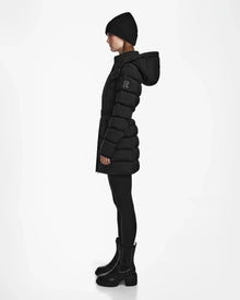 A woman stands in profile wearing the Rudsak FRIDA M - L124524 Black womens mid-length down jacket with removable hood, a black beanie, leggings, and ankle boots against a plain white background.