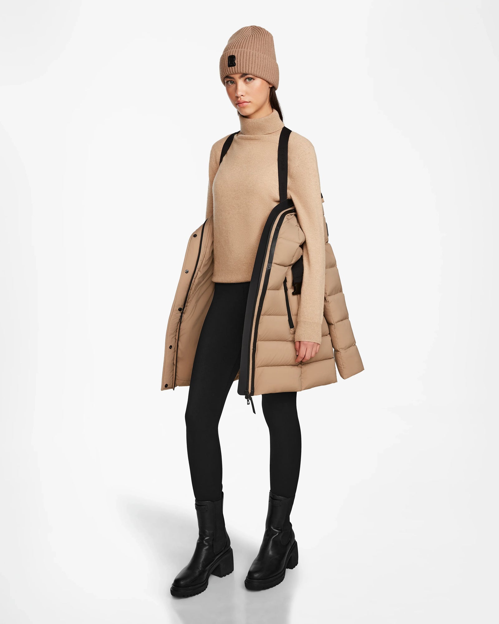 A woman in a beige FRIDA M - L124524 Sand jacket by Rudsak with a removable hood draped over one shoulder stands against a plain white background, wearing a beige beanie, turtleneck, black pants, and black boots.