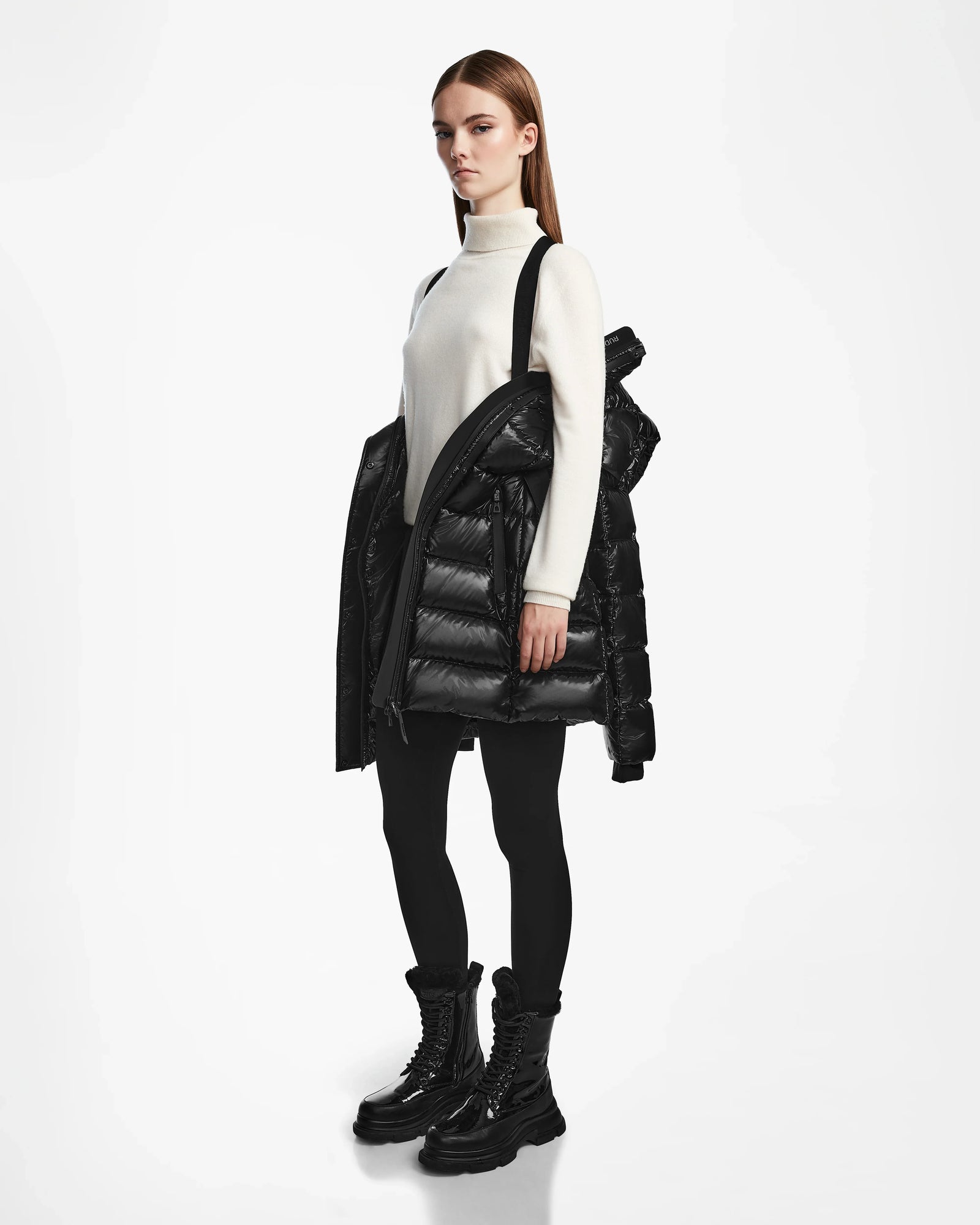 A woman stands against a white background wearing a white turtleneck, black leggings, and black boots, holding the Rudsak FRIDA S - L124526 Black quilted down jacket with removable hood draped over her arm.