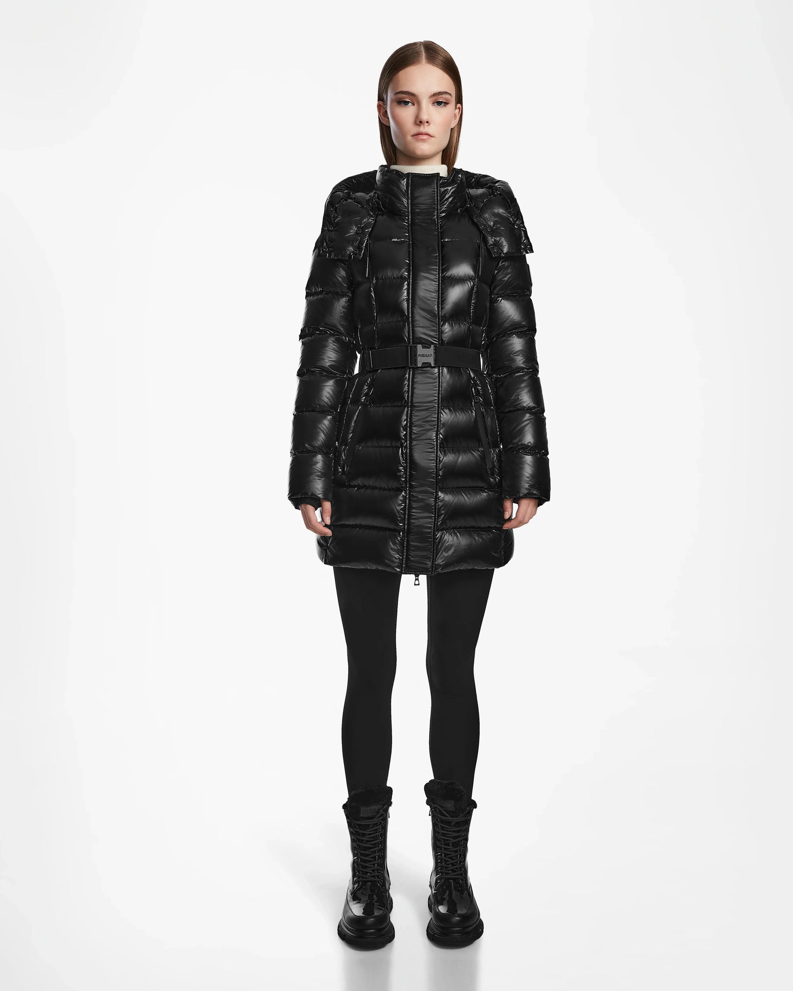 A woman faces forward against a plain white background, wearing the Rudsak FRIDA S - L124526 Black womens mid-length down jacket with a belted waist, paired with black leggings and black lace-up boots.