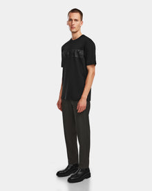 A man stands against a plain background wearing the Rudsak GIANNI - 6524535 Black t-shirt, paired with dark gray pants and black boots. He faces forward with a neutral expression, giving the modern outfit a relaxed vibe.