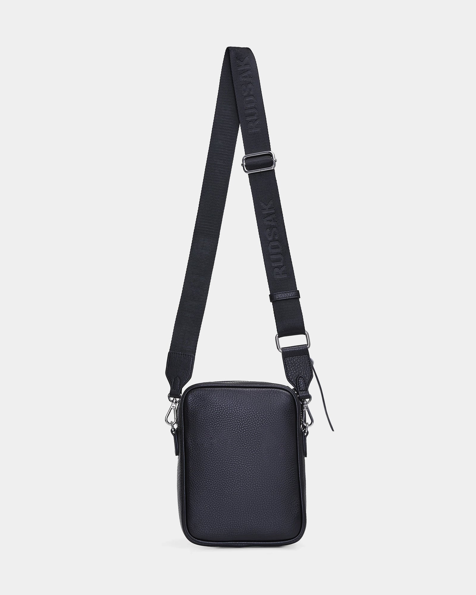 The Rudsak HUBERT - M324500 Black is a small, rectangular crossbody bag with a textured black surface, silver hardware, and an adjustable strap with embossed RUDSAK branding, shown against a plain white background.