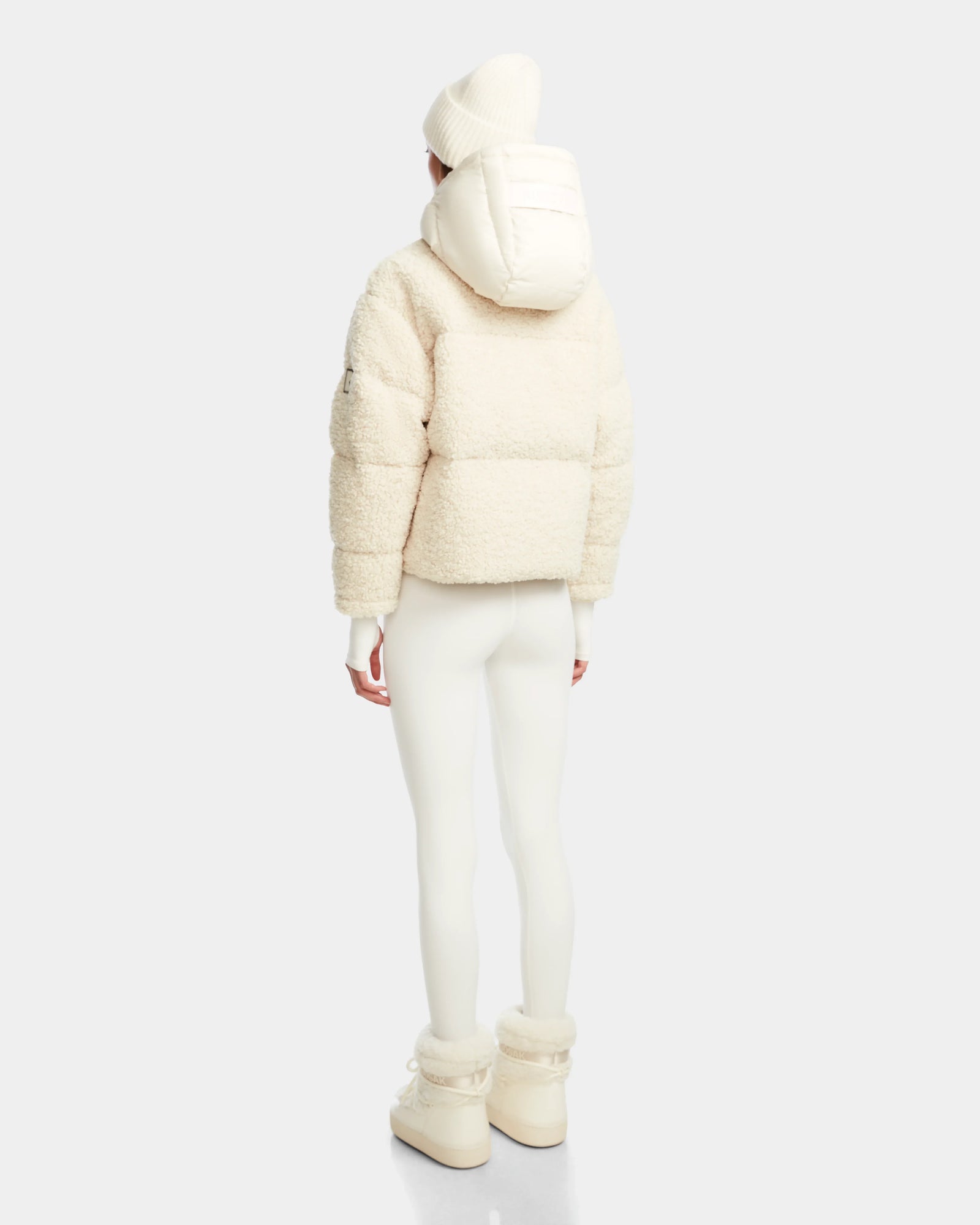A person wearing the Rudsak IKSA - 8124706 Iceland RDS-certified down jacket with hood, white beanie, leggings, and boots stands facing away against a plain light background.
