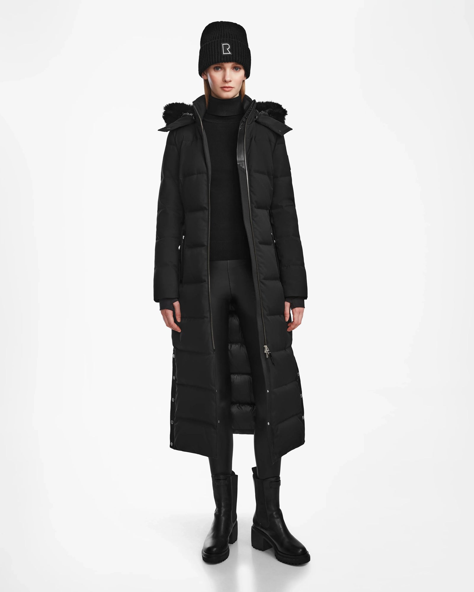 A person wearing the Rudsak IZABELLA - L124541 Black women's long down coat, with a beanie, turtleneck sweater, black pants, and boots stands facing forward against a plain white background.
