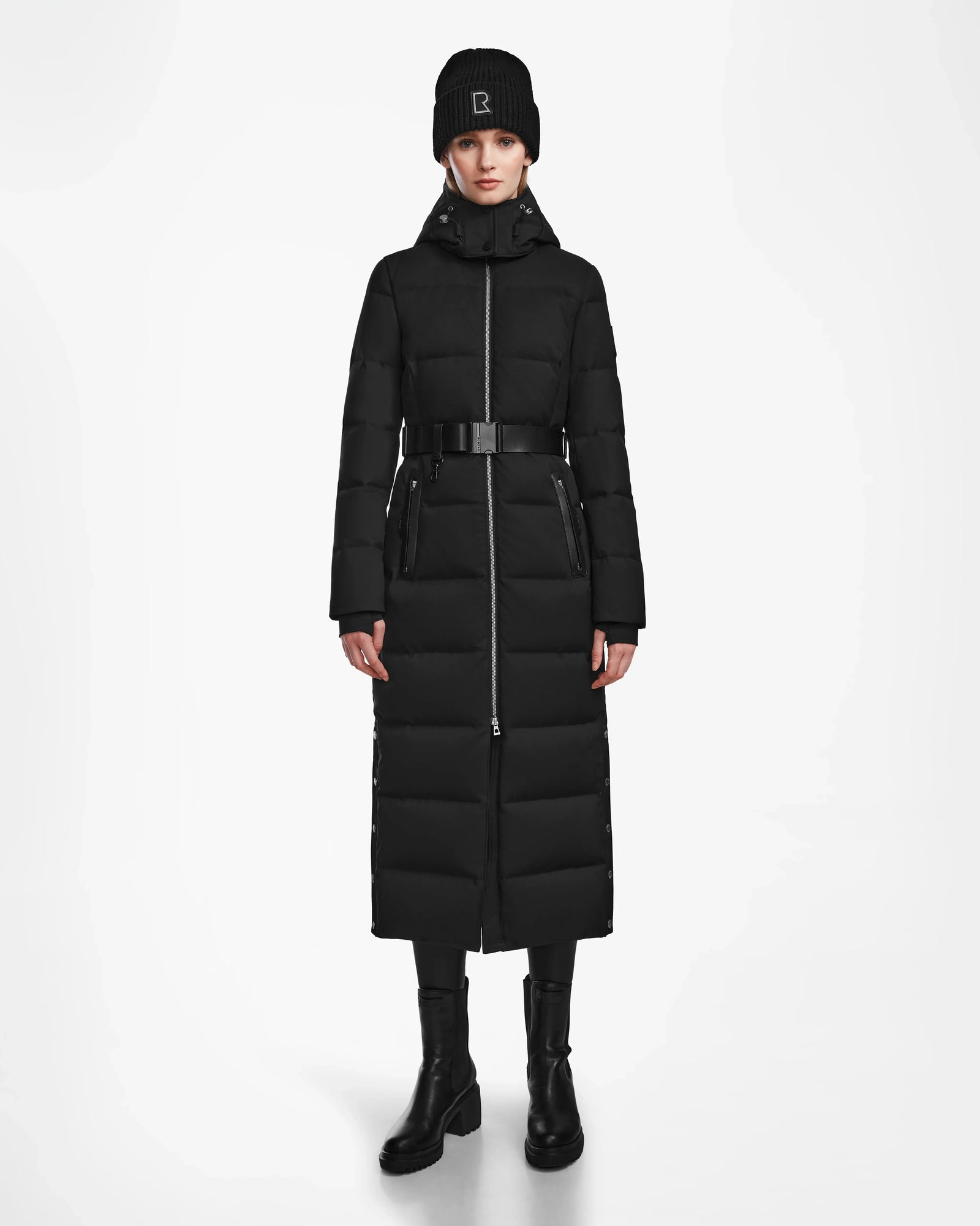 A person stands facing forward wearing the Rudsak IZABELLA - L124541 Black women's long down coat, paired with a black beanie, black pants, and ankle boots against a plain white background.