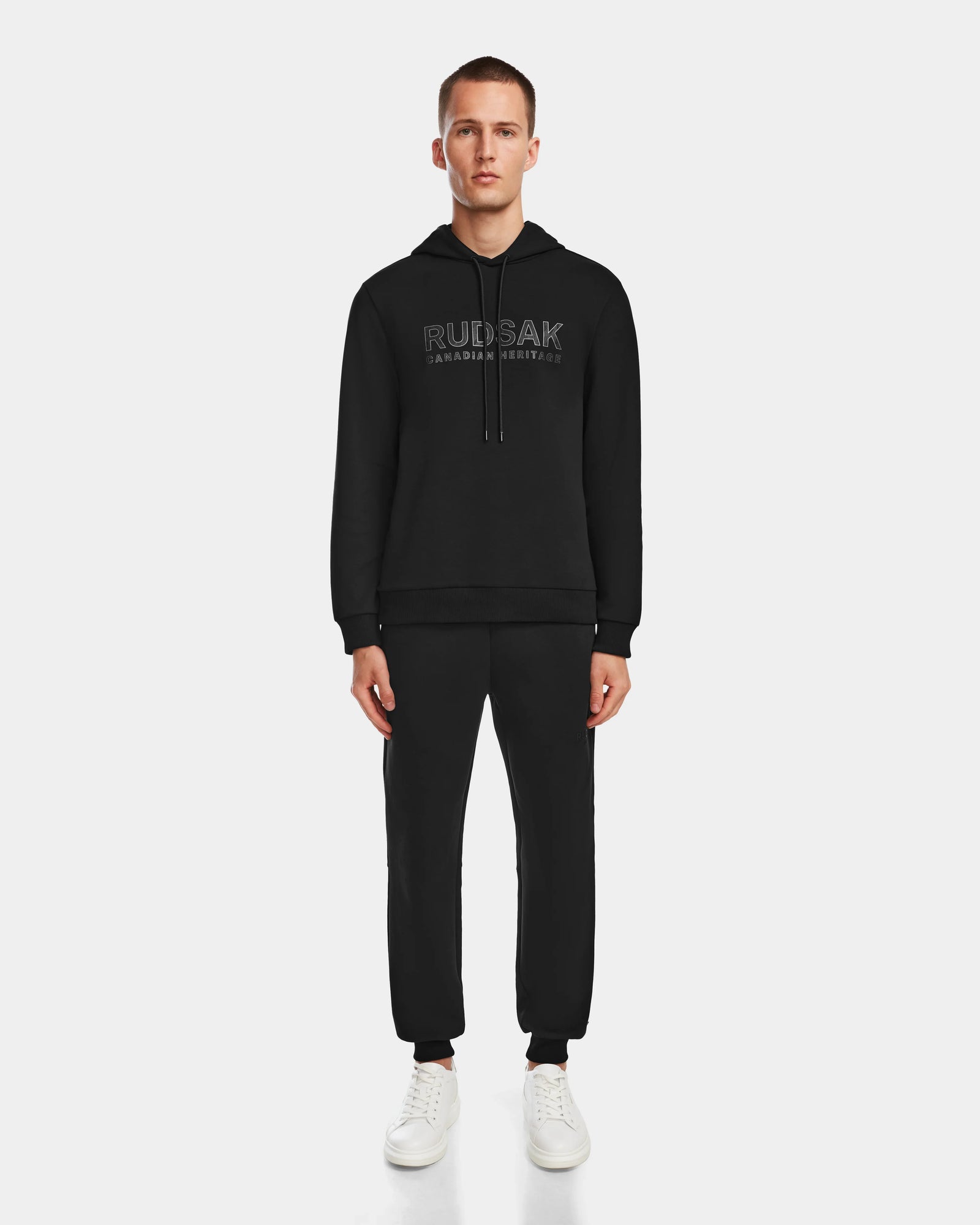 A man faces forward against a white background, wearing the black Rudsak JARED - 6524533 hoodie and matching jogger pants, paired with white sneakers.
