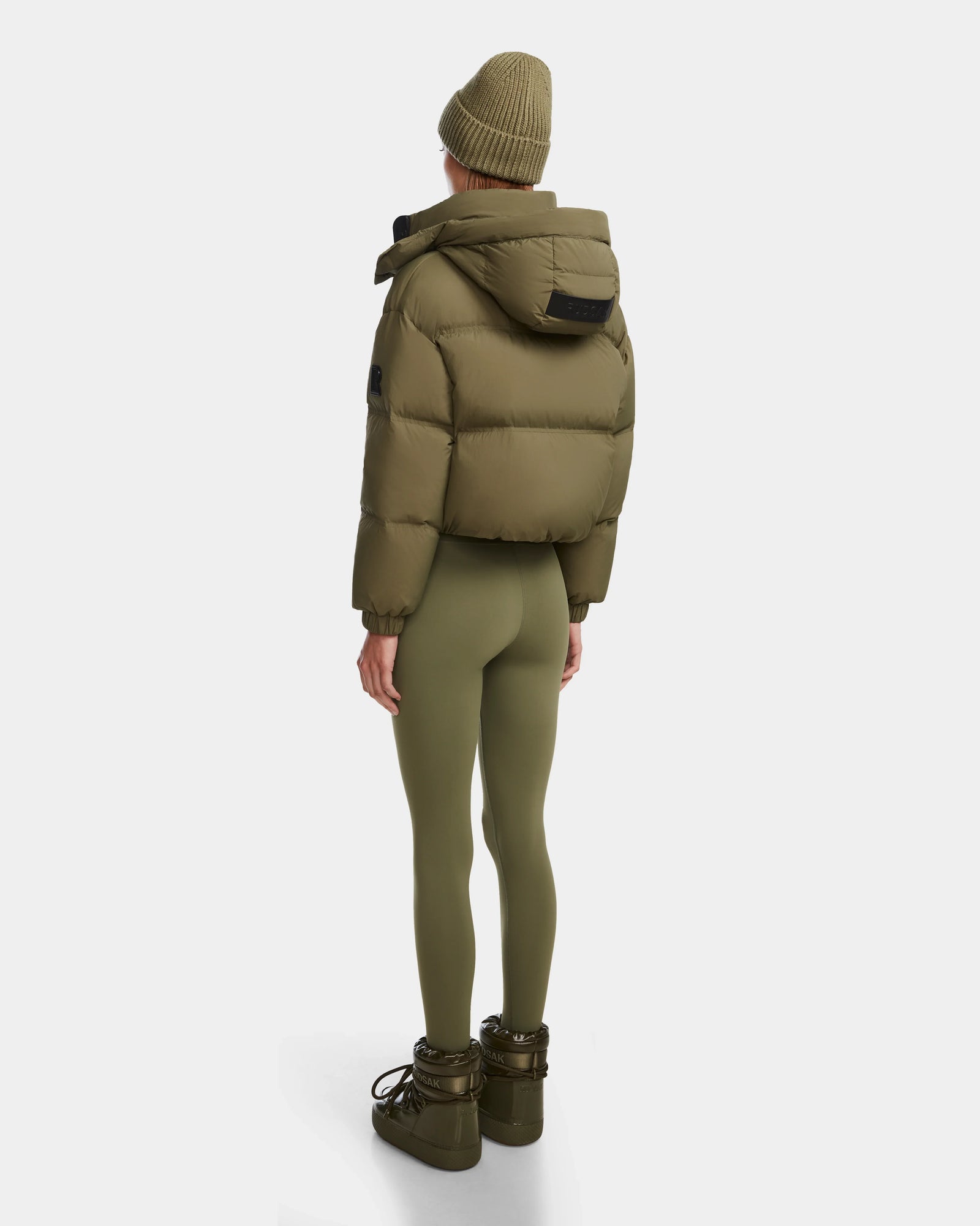 A person stands facing away, wearing the Rudsak KARLEE - 8124721 cropped down puffer in Loden Green with a matching beanie, fitted leggings, and boots. The quilted jacket pops against a plain white background.